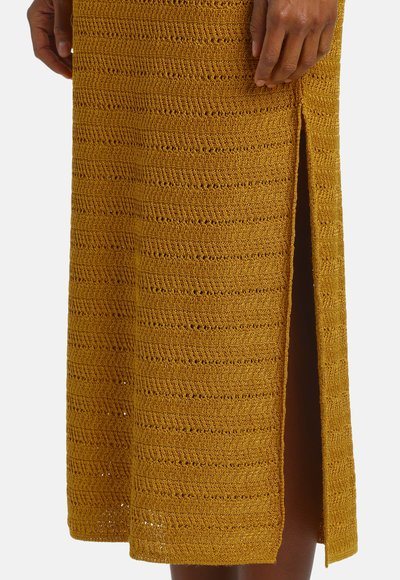 Jupe en maille jaune moutarde avec motif texturé horizontal et fente latérale, portée par une personne les mains posées près de la taille.