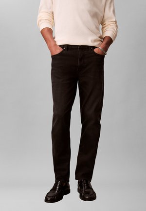 Mann trägt schwarze Jeans, schwarze Lederloafer, cremefarbenes Langarmhemd, Hände in den Taschen, steht vor einem einfarbigen grauen Hintergrund.