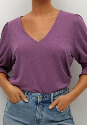 Fioletowa koszulka z dekoltem w serek i bufkami na rękawach, wykonana z miękkiej tkaniny. Połączona z jasnoniebieskimi szortami z wysokim stanem z denimu. Dwie dłonie w kieszeniach.