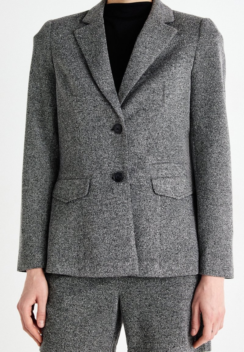 Grå texturerad kavaj med slagkrage, två framfickor och två svarta knappar. Bärs över en svart topp och matchande byxor.