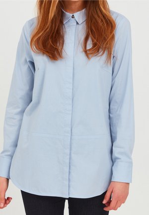 Kvinde iført en lyseblå langærmet skjorte med knapper og krave, kombineret med mørke bukser, stående foran en hvid baggrund.