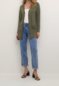 Olivově zelený kardigan s výstřihem do V, dlouhými rukávy a dvěma předními kapsami; doplněný světle modrými zúženými džíny vyhrnutými na kotnících a černými sandály na blokovém podpatku.