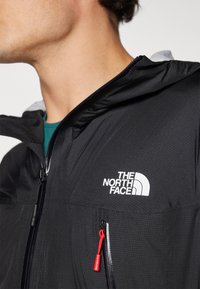 Veste légère noire avec poches zippées, arborant un logo blanc et une tirette rouge. Tissu texturé avec une capuche.
