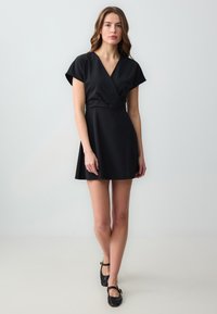 Schwarzes Wickelkleid mit kurzen Ärmeln, V-Ausschnitt und ausgestellt Rock. Glattes Gewebe, minimalistisches Design. Schwarze Ballerinas mit Knöchelriemen.