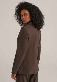 Blazer marrone su misura con rever a punta, apertura posteriore singola e una texture liscia, modellato con una silhouette aderente e maniche lunghe.