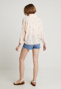 Kobieta z krótkimi brązowymi włosami, ubrana w luźną bladoróżową koszulę z małymi zielonymi printami, dżinsowe szorty i białe sandały, stojąca na białym tle.