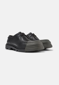 Scarpe in pelle nera con punta arrotondata e cappuccio in gomma, caratterizzate da una suola resistente e dettagli in cucitura minimi. I lacci sono posizionati centralmente.