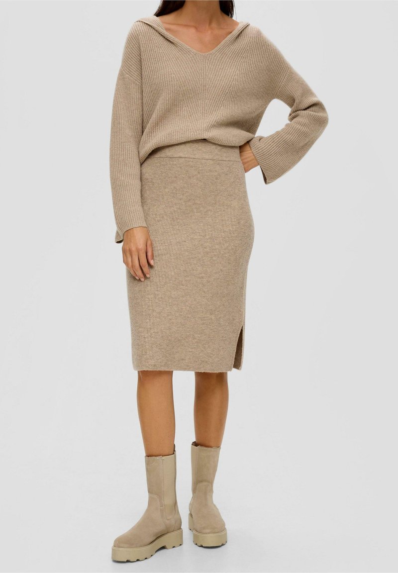 Femme portant une jupe beige en maille côtelée avec une fente latérale, un pull ample à col en V assorti, et des bottes épaisses beiges mi-mollet sur fond gris.