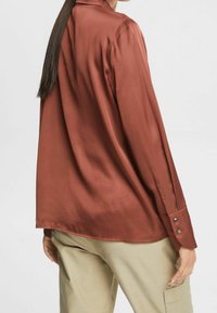 Vrouw met lang donker haar, gekleed in een roestkleurige satijnen blouse en beige cargo broek, van de achterkant gezien tegen een effen achtergrond.