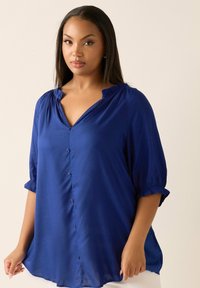 Blå knäppblus med avslappnad passform, volangkrage och korta ärmar. Tillverkad av lätt vikt tyg med en slät textur.