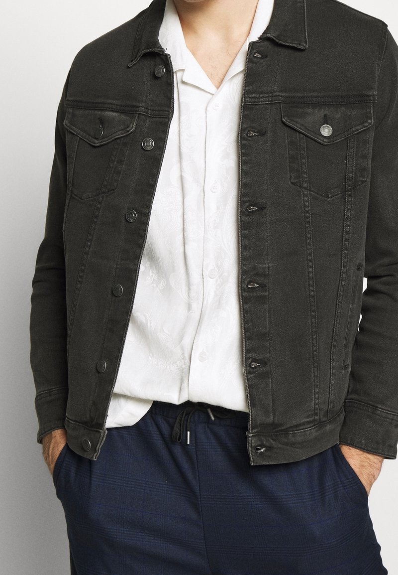 Tmavě modrá jeanová bunda s dvěma prsními kapsami, stříbrnými knoflíky a opotřebovanými detaily, nošená přes bílou košili, spárována s námořnicky kostkovanými kalhotami.