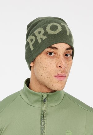 Olivgrüner Strickbeanie mit deutlich sichtbarem "PROTEST"-Schriftzug. Glatte Textur, enge Passform und weicher Stoff. Getragen mit einem passenden hochkragen Oberteil.
