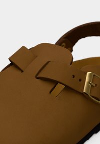 Καφέ δερμάτινο σανδάλι με χρυσαφένια αγκράφα και ρυθμιζόμενο λουράκι σε λευκό φόντο.