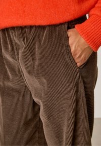 Rjave kordne hlače z vertikalno rebrasto teksturo, ki imajo žep in so oblečene z svetlo oranžno puloverjem, ki prikazuje mehko pletenino.