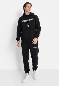 Set di felpa con cappuccio nero e pantaloni della tuta, caratterizzato da una grande stampa del logo bianco, cappuccio con cordino, tasca frontale e polsini a costine. Sneakers bianche.