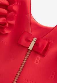 Czerwona odzież o teksturowanej tkaninie, z falbaniastymi akcentami i centralnym złotym zamkiem. Logo "BAKER" delikatnie wytłoczone w materiale.