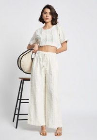Weißes Crop-Top mit strukturierten Streifen, kombiniert mit weit geschnittenen Hosen; das Model hält eine runde, geflochtene Tasche. Klare, genietete Sandalen runden das Outfit ab.