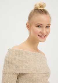 Maglione lavorato a costine beige scivolato sulle spalle, con una consistenza morbida, caratterizzato da una scollatura ampia e maniche aderenti, esposto su uno sfondo neutro.