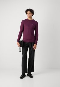 Maglione viola a girocollo realizzato in un tessuto liscio. Abbinato a pantaloni neri a gamba larga e scarpe nere lucide. Porta una piccola borsa nera.