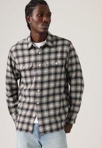 Άνδρας με πλεξούδες, που φοράει μαύρο και άσπρο καρό πουκάμισο πάνω από λευκό μπλουζάκι και ανοιχτόχρωμο μπλε τζιν, κοιτάζοντας προς το πλάι.