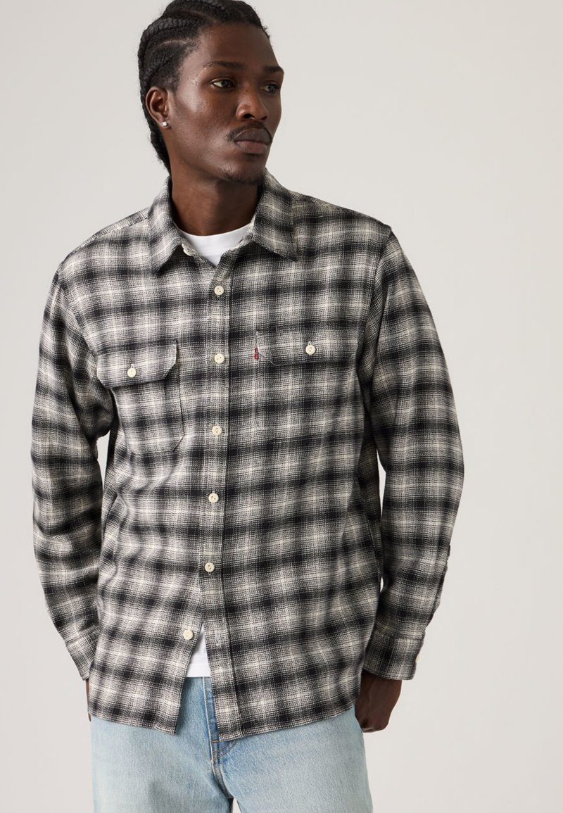 Homme aux cheveux tressés portant une chemise à carreaux noire et blanche boutonnée sur un t-shirt blanc et un jean bleu clair, regardant sur le côté.