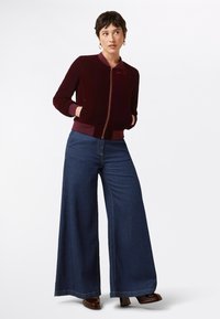 Giacca bomber in velluto bordeaux con zip frontale, polsini a costine e due tasche laterali, abbinata a jeans in denim scuro a gamba larga.