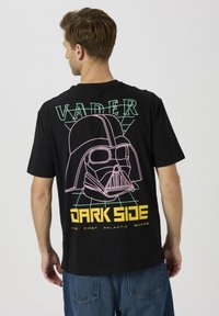Svart bomulls-T-skjorte med et bakgrafikk av en stilisert Darth Vader-hjelm i rosa og grønn, med teksten "VADER" og "DARK SIDE".