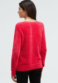 Frau mit langen dunklen Haaren, die einen leuchtend roten Chenille-Pullover mit Text auf dem linken Ärmel trägt, steht vor einem schlichten hellen Hintergrund.