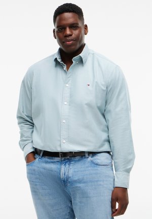 Mand med kort krøllet hår iført lyseblå skjorte med knapper og lyseblå jeans, stående med den ene hånd i lommen mod hvid baggrund.