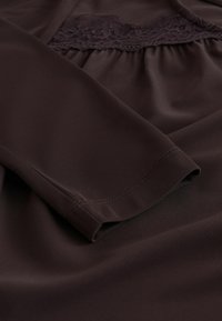 Haut en tissu marron avec un panneau en dentelle texturée près de la poitrine, manches longues et coupe décontractée avec des coutures piquées.