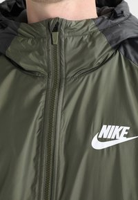 Olivgrön jacka med svart huva, dragkedjestängning och vit Nike-logotyp på vänster bröst. Slätt, lätt material och texturerade detaljer.