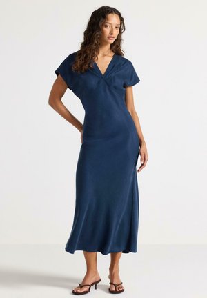 Kvinde med krøllet hår iført en mørkeblå midi-kjole med korte ærmer og V-hals, poserer med den ene hånd på hoften.