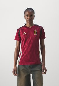 Sarkans futbols kreklis ar dimanta rakstu, melniem un zelta akcentiem, ar V-veida kaklu un īsām piedurknēm, ar Beļģijas karoga emblēmu uz krūtīm.