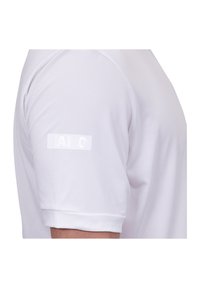Camicia bianca a maniche corte realizzata in tessuto elasticizzato, con una texture liscia e un logo stampato sulla manica sinistra. Dettaglio cucitura lungo la spalla.