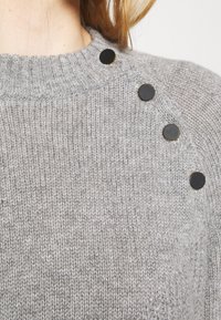 Maglione grigio a maglia con scollatura a costine, texture liscia e cinque bottoni neri lungo una spalla.
