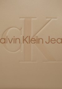 Beige tekstureret materiale med præget "Calvin Klein Jeans" logo. Bemærkelsesværdige træk inkluderer en glat overflade og subtil glans.