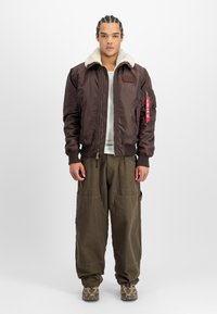 Chaqueta bomber marrón con cuello de piel sintética, cierre de cremallera y puños acanalados. Presenta un parche en el pecho y una etiqueta en la manga.