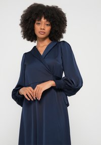 Marineblå omslagskjole med lange puffermede ermer, glatt stoff, dyp V-utringning og innsvingt midje. Har detaljer med sidestropp og en flytende skjørt.