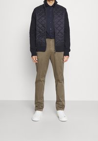 Giacca trapuntata blu navy con maniche in maglia, indossata sopra una camicia blu navy con pois. I chinos color sabbia e le sneakers bianche completano l'outfit.