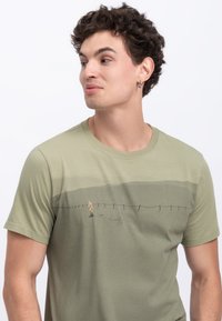 T-shirt verde oliva realizzato in morbido cotone, con un delicato design paesaggistico che raffigura una figura che cammina lungo una recinzione, maniche corte e collo rotondo.