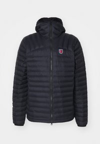 Sötét tengerészkék, párnázott dzseki cipzáros elülső résszel, kapucnival és mellkasi logóval. Vízszintes steppelt részek és elasztikus mandzsetta jellemzi. Simább textúra.