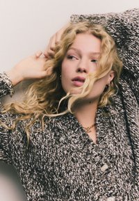 Biezs dzijas cimdi pelēkā un baltā krāsā, ar dziļu V-veida izgriezumu un lielām pogām. Raksturīga teksturēta auduma un slāņveida dizaina. Redzama zelta ķēdes kaklarota.