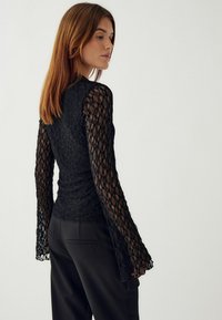 Čierny čipkový top s dlhými, rozšírenými rukávmi a priliehavým telom. Textúra má diamantový vzor a je doplnená vysokými čiernymi nohavicami.