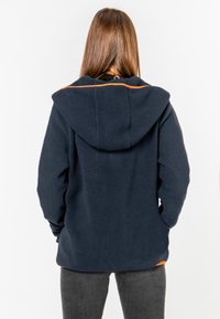 Tummansininen fleece-takki hupulla, jossa on oranssi yksityiskohta selän yläosassa, teksturoitu pinta ja rento istuvuus.