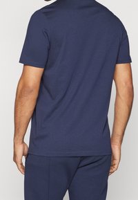 Mężczyzna ubrany w gładki, granatowy t-shirt z krótkim rękawem i pasujące granatowe spodnie, pokazany od tyłu na jednolitym tle.