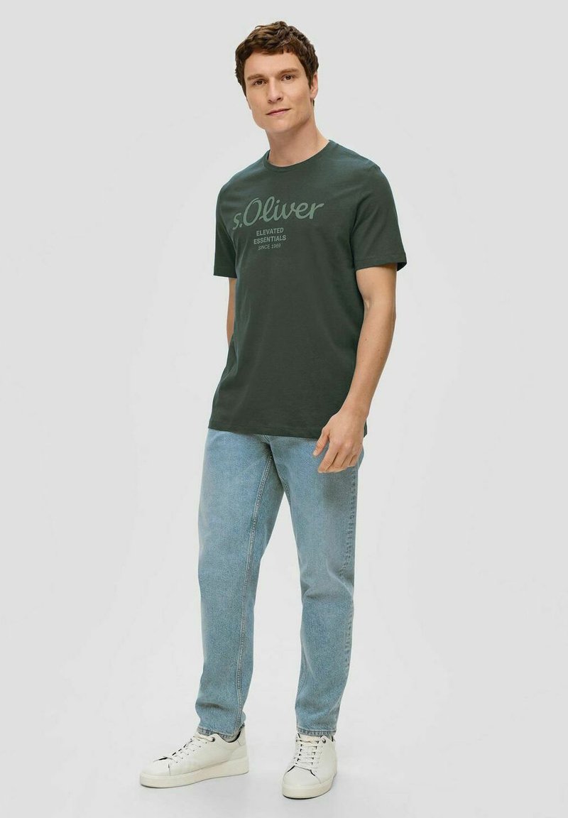 Dunkelgrünes Baumwoll-T-Shirt mit kurzen Ärmeln, das ein dezentes Drucklogo-Design aufweist. Kombiniert mit hellblauen Jeans und weißen Sneakers.