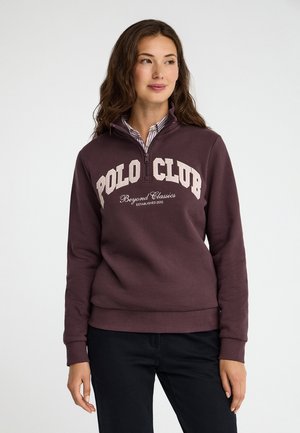 Mujer con sudadera granate con cremallera parcial y texto "Polo Club Beyond Classics", combinada con pantalones negros, de pie sobre fondo blanco.