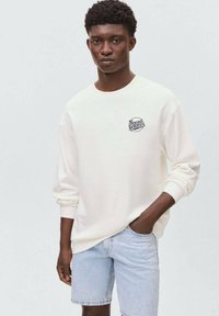Sweatshirt blanc avec des poignets côtelés, arborant un logo bleu "Beyond Horizon" sur la poitrine gauche, associé à un short en denim clair.