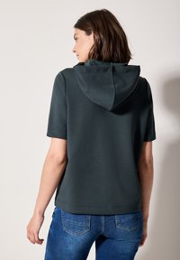 Sudadera corta de manga corta en verde oscuro con capucha ajustable, textura suave y corte relajado, combinada con jeans de color azul claro.
