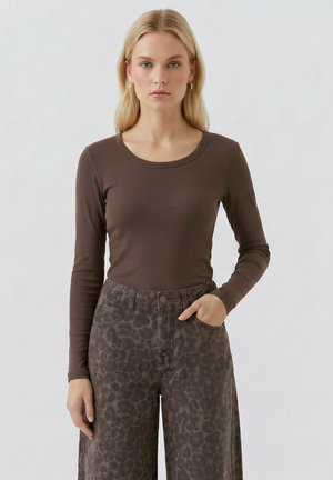 Frau mit langen blonden Haaren, die ein braunes Langarmoberteil und hoch taillierte, dunkle Hose mit Leopardenmuster trägt, steht vor einem schlichten Hintergrund.